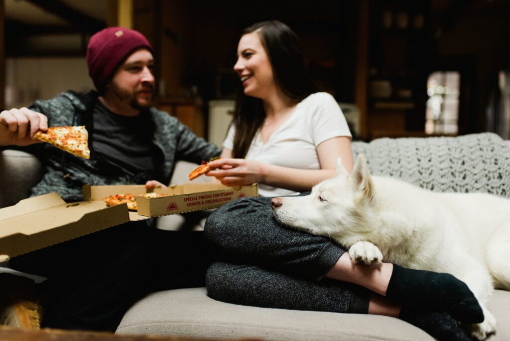 a man and woman on a couch eating pizza out of a pizza box with a dog laying on the woman's legs