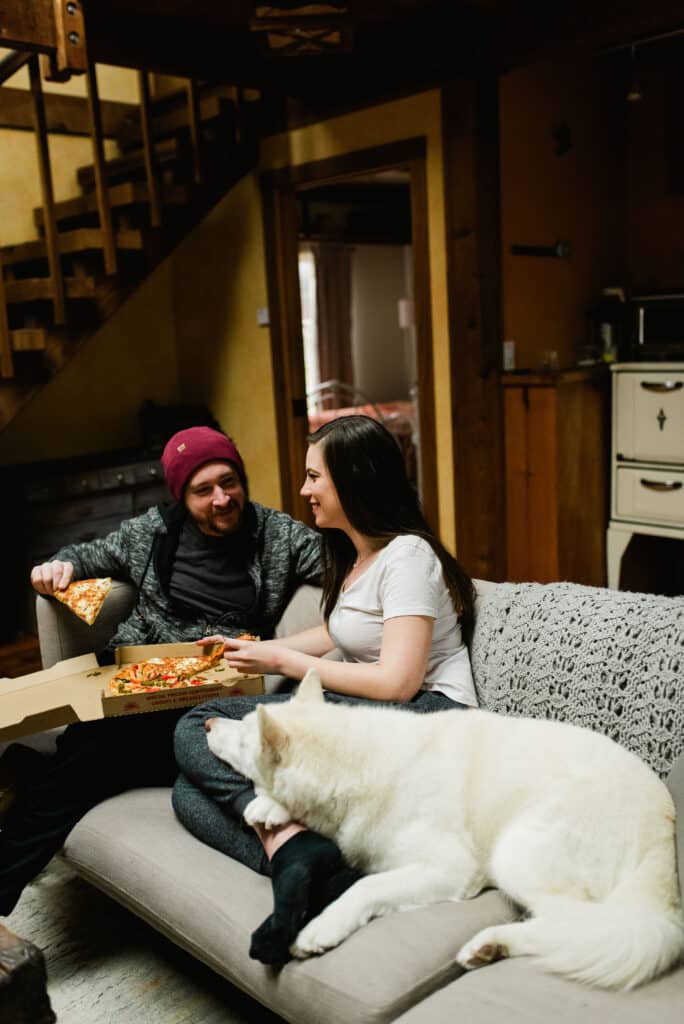 a man and a woman sitting on a couch eating pizza out of a pizza box with a big white dog laying on the woman's legs